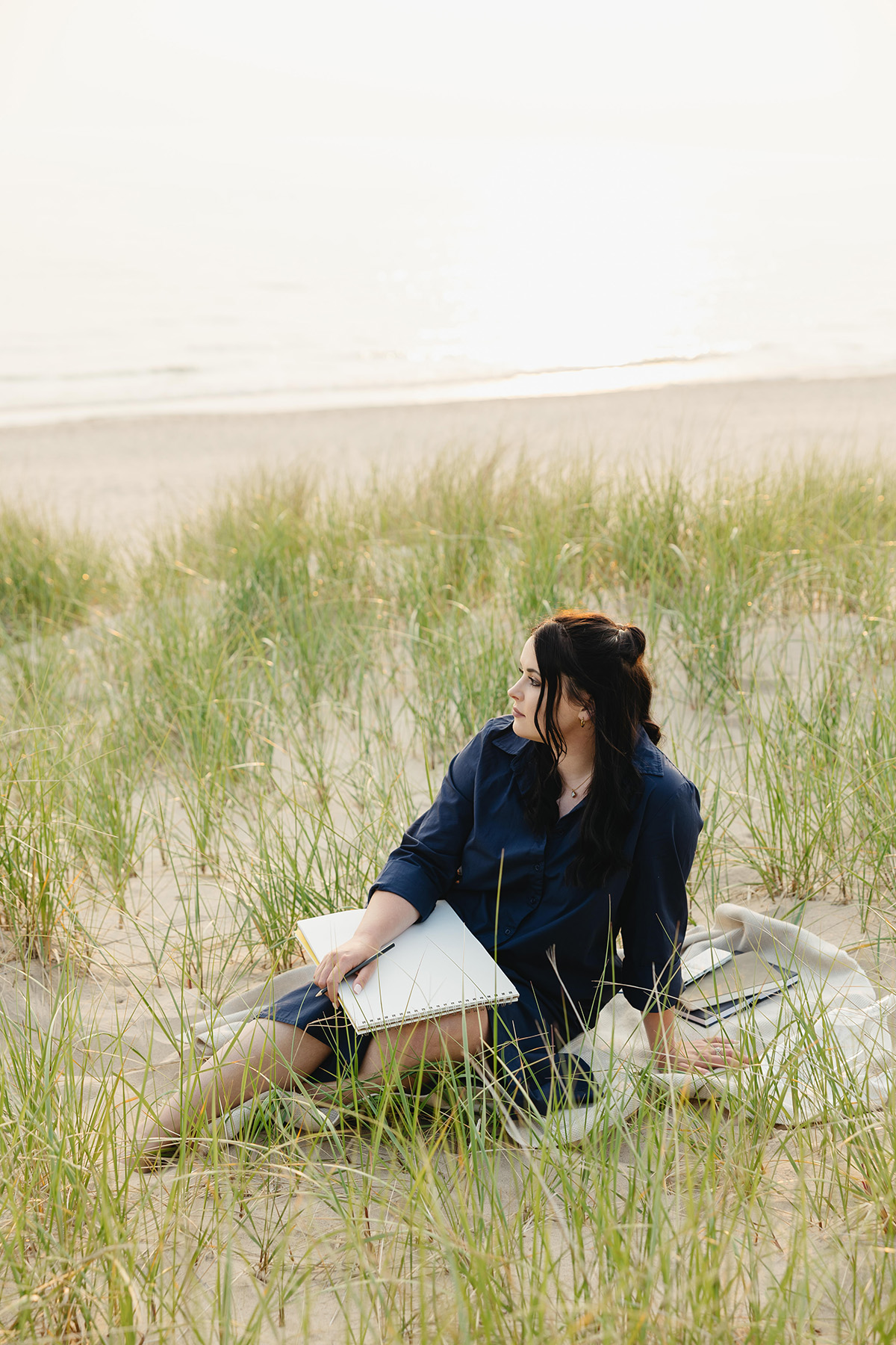 woman sitting in the sand and beach grass looking out to Lake Michigan at sunset